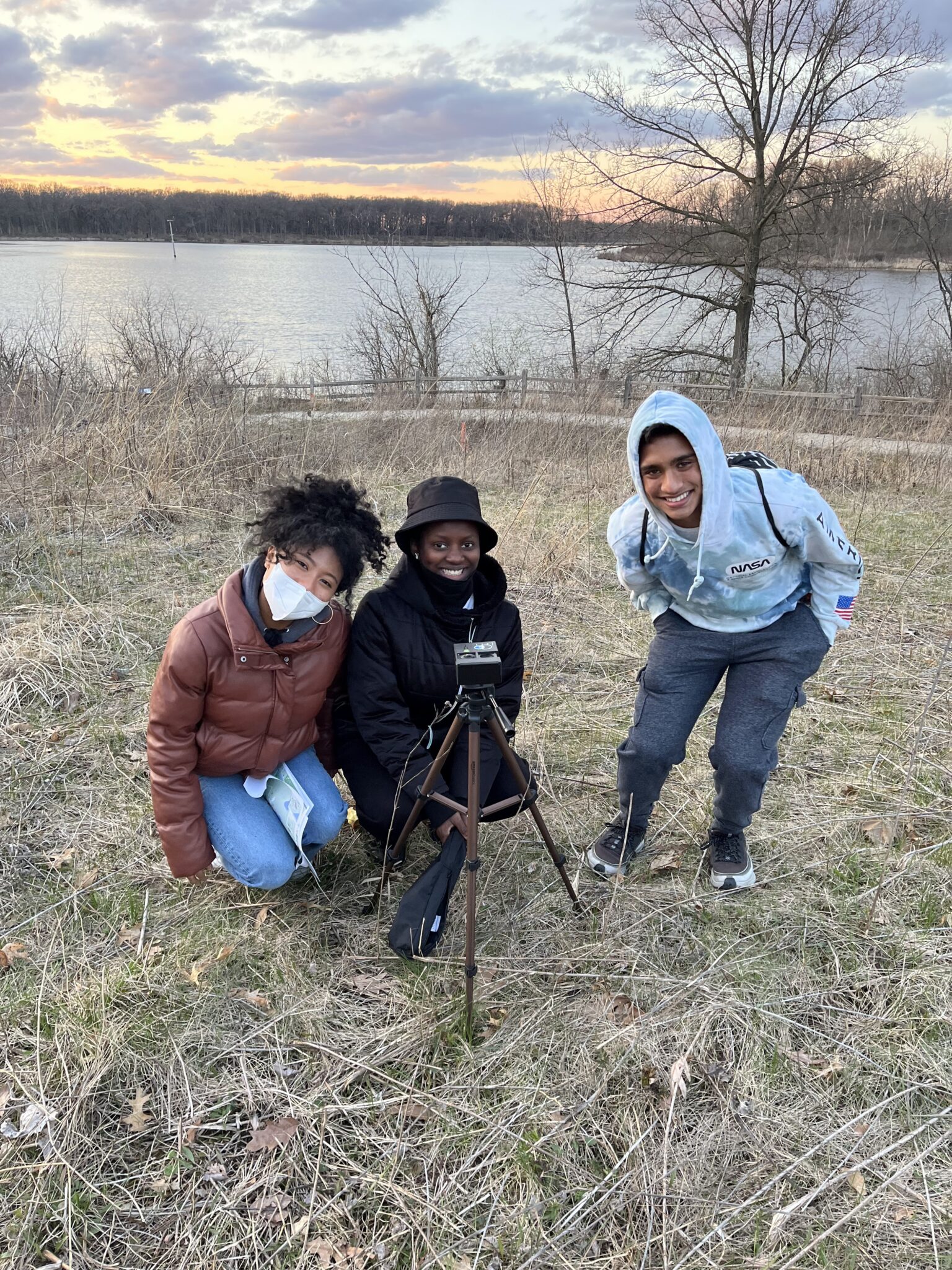 Three students pose for a picture in a field in front of a lake.
